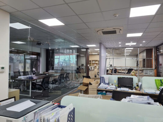 Hình ảnh một văn phòng hiện đại với thiết kế mở, sử dụng vách kính lớn ngăn khu làm việc và phòng họp, bàn ghép từng không gian nhỏ và ánh sáng tự nhiên từ cửa sổ lớn.